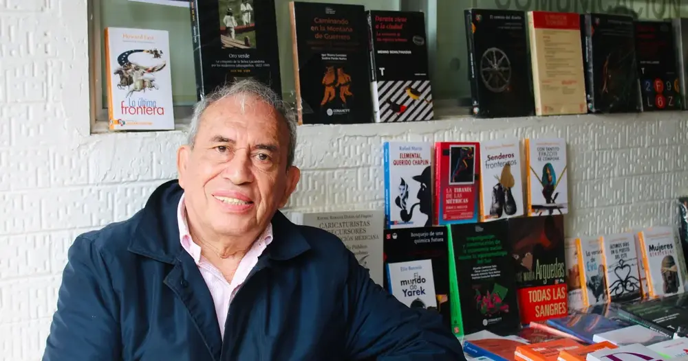 Um homem sorridente está sentado perto de uma banca de livros com diversos títulos publicados, exibindo sua coleção.